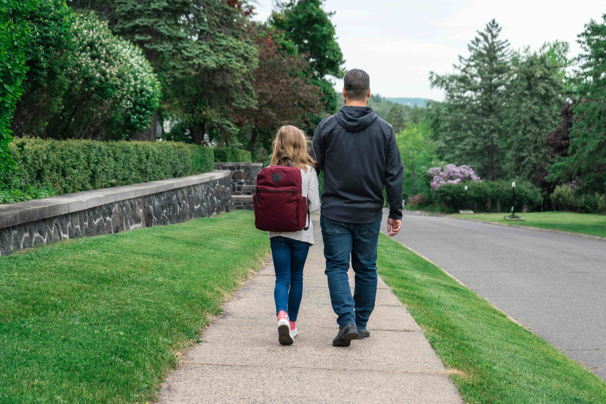 A dad and daughter walking down the street. The daughter is wearing a burgundy Standard Backpack by Duluth Pack for back to school.  