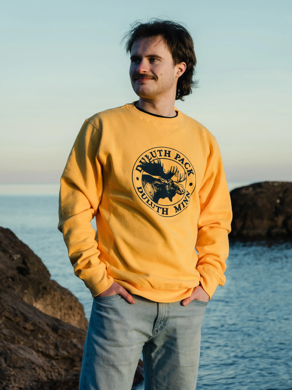 Man wearing a yellow Duluth Pack Crewneck with a lake in the background