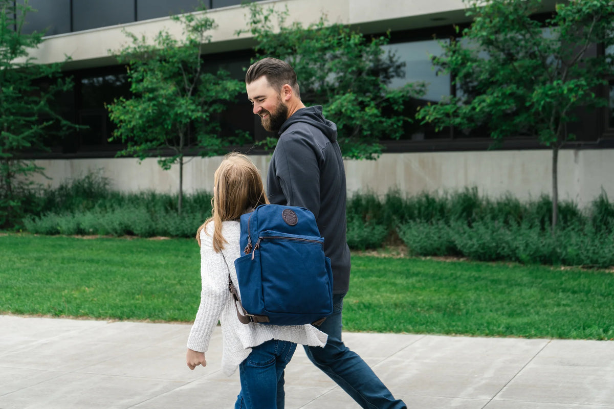 A dad and daughter walking to school with a Duluth Pack New Standard Backpack in Navy with a school in the background. 
