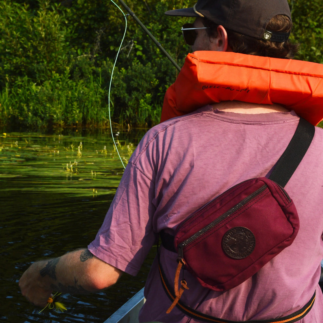 A man wearing a duluth pack terraventure fanny pack in burgundy. He is fly fishing in the boundary waters canoe area.