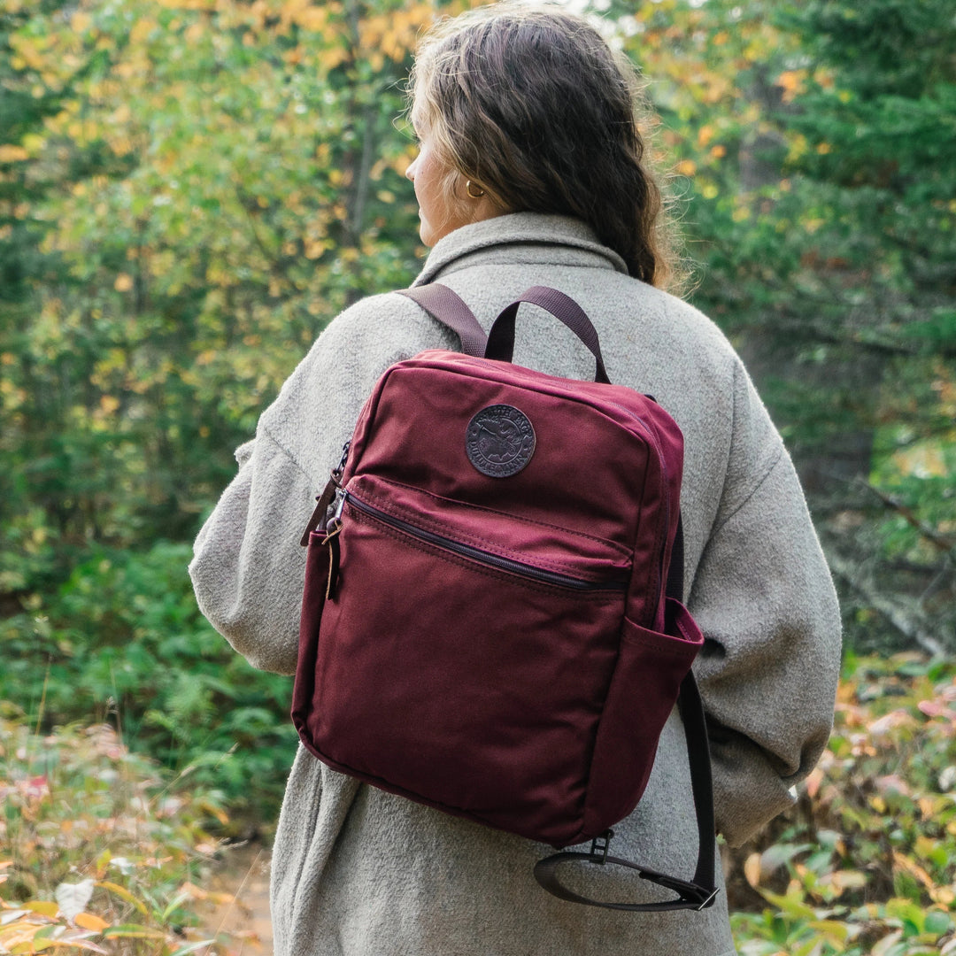 A woman wearing a duluth pack standard backpack in burgundy with a fall forest in the background 